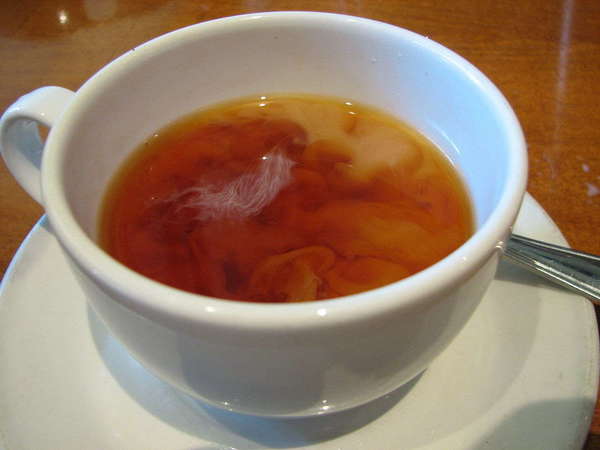 A white cup on a white saucer, filled with reddish-brown tea with swirling cloud shapes from added milk