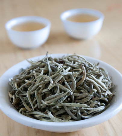 Loose leaf tea consisting of silver-green tips in a white dish, two blurry cups of pale yellow tea in background