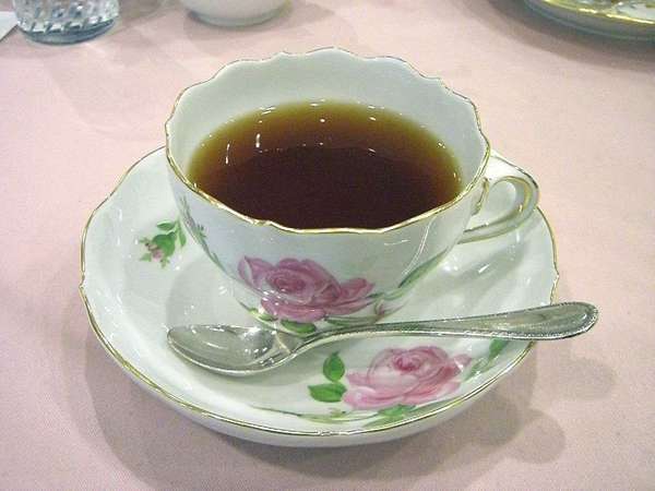 Victorian rose-patterned tea cup filled with black tea, with matching saucer and shiny spoon, on a pink background
