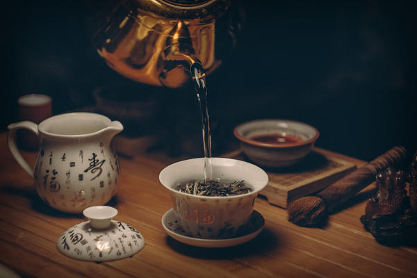 A metal tea kettle pouring hot water into a gaiwan with loose-leaf tea, other teaware surrounding on table